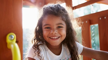 Young Hispanic girl playing on a climbing frame in a playground smiling to camera, backlit, close up