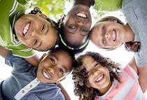A multi-ethnic group of elementary age children are huddled together, standing in a circle with their heads in the center looking down at the camera. They are smiling and looking at the camera.
