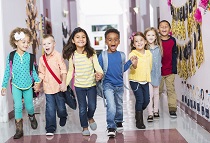 A multi-ethnic group of seven children holding hands, running down their school hallway, laughing and shouting, looking at the camera.  The little boys and girls are kindergarten or preschool age, 4 to 6 years.